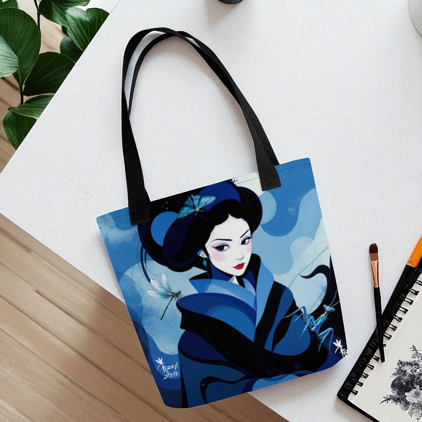 Bolso tote de geisha azul visto desde arriba, tumbado en mesa blanca con pincel y cuaderno artístico al lado.