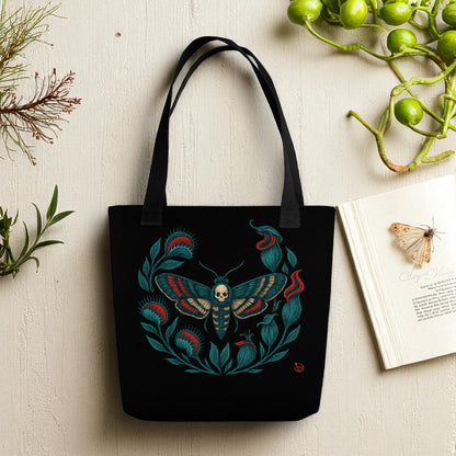 Bolso tote negro con diseño de polilla calavera y hojas, vista frontal con asas negras sobre mesa.