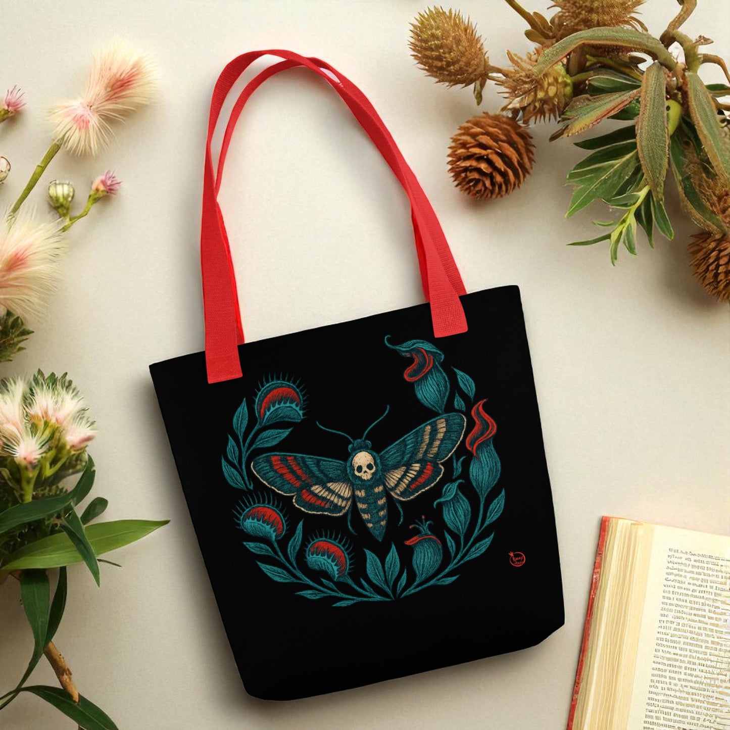 Bolso tote negro con polilla calavera y asas rojas en escena decorativa con plantas.