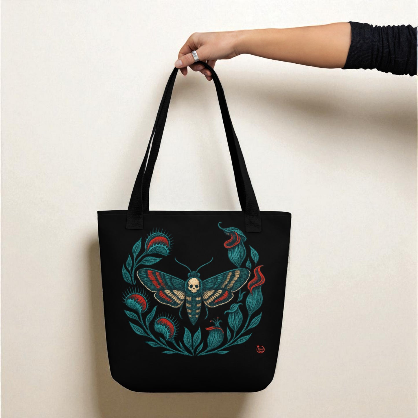 Mano sosteniendo bolso tote negro con polilla calavera, asas negras.