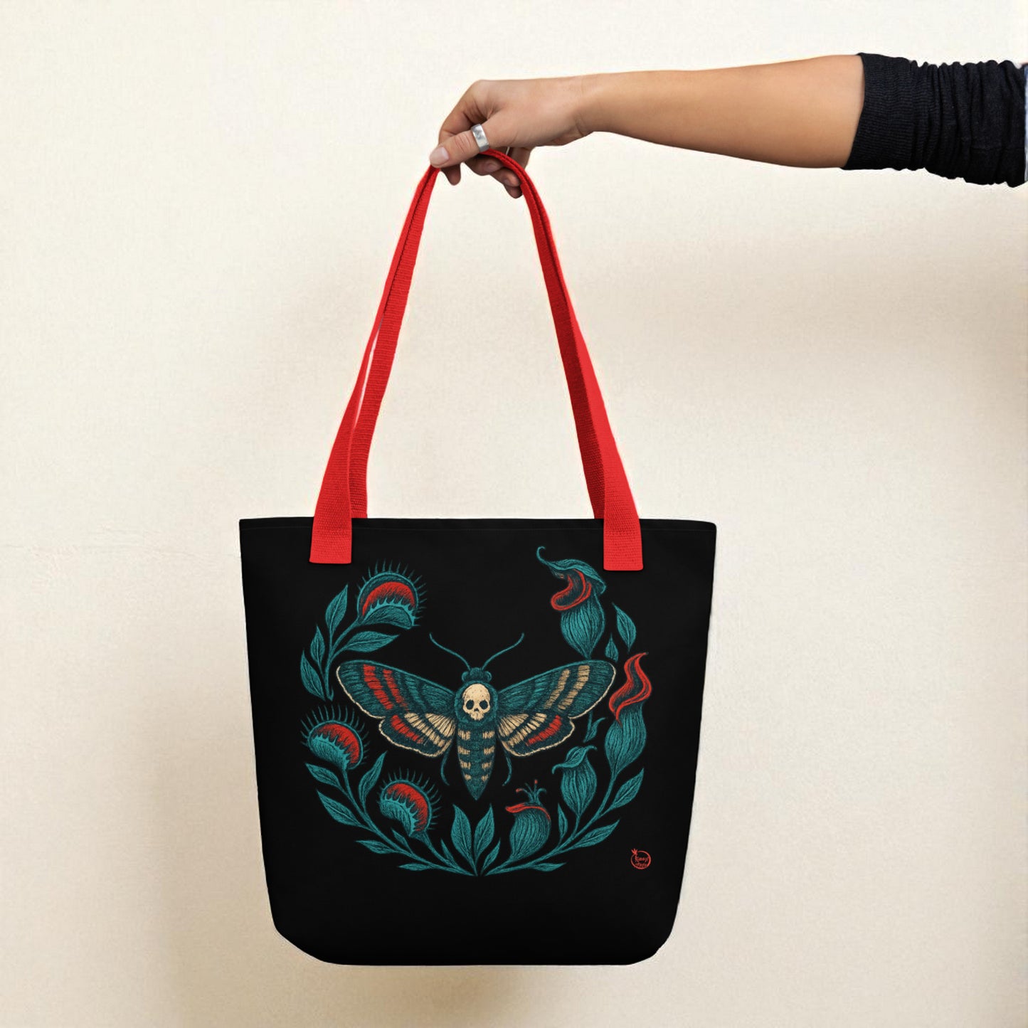 Mano sosteniendo bolso tote negro con polilla calavera, asas rojas.