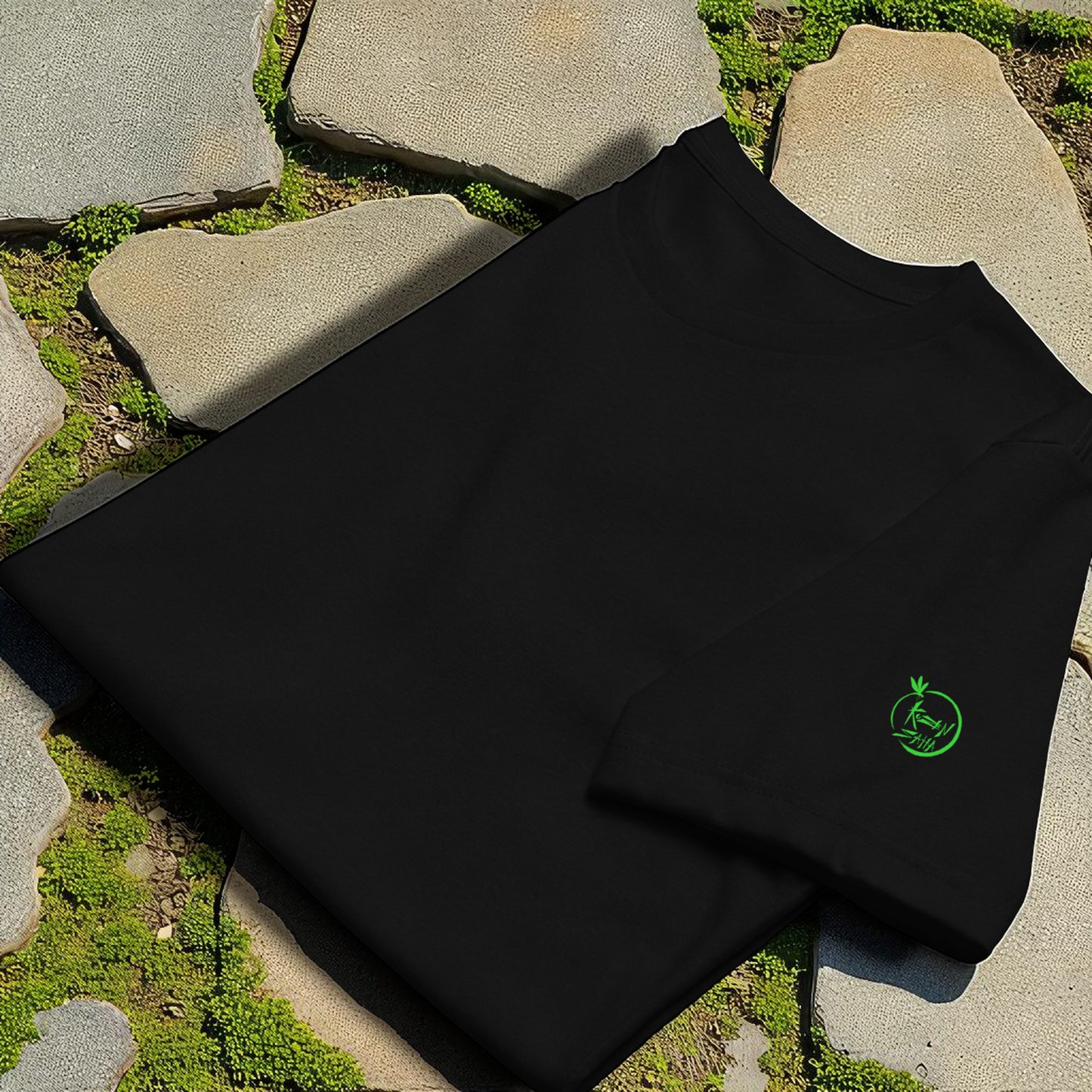 Camiseta negra doblada sobre piedras,detalle de logo Kuzen Saha con libéliula en verde en manga izquierda