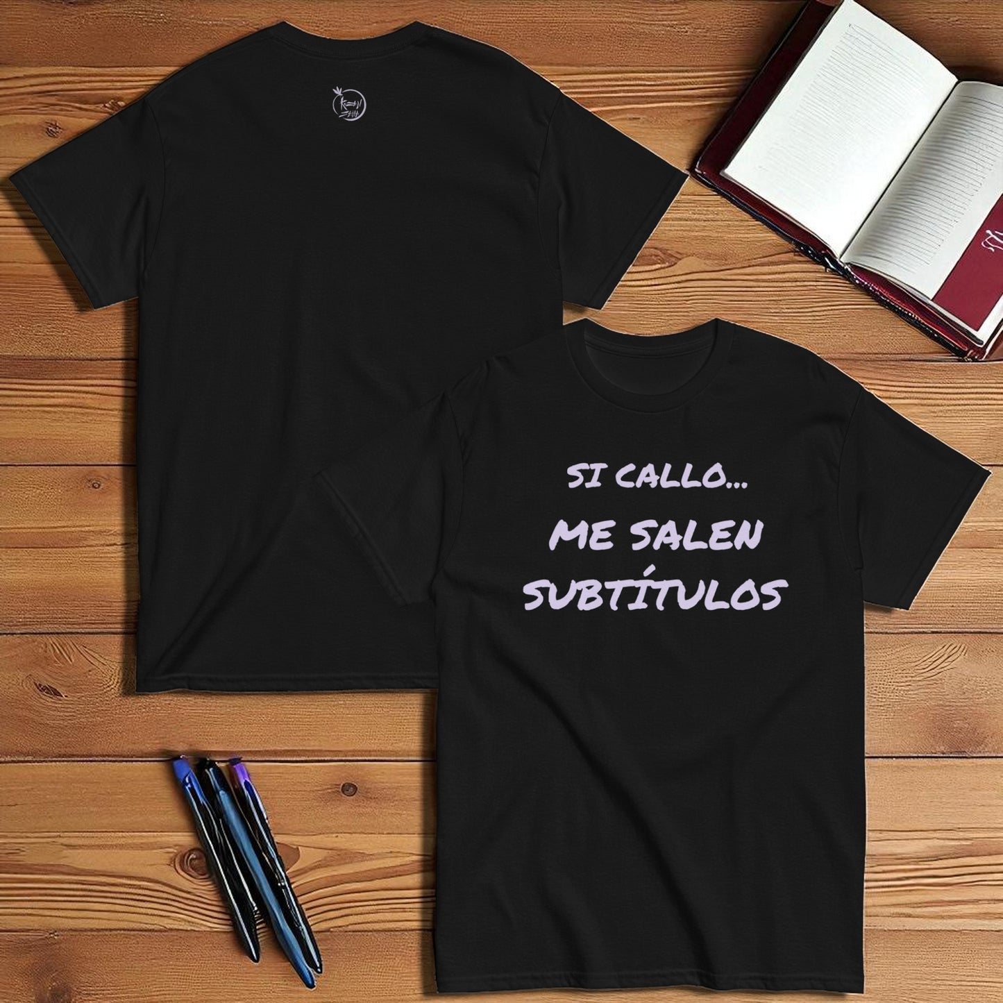 Camiseta negra unisex con frase Si callo me salen subtítulos en plano cenital con libreta y lápiz