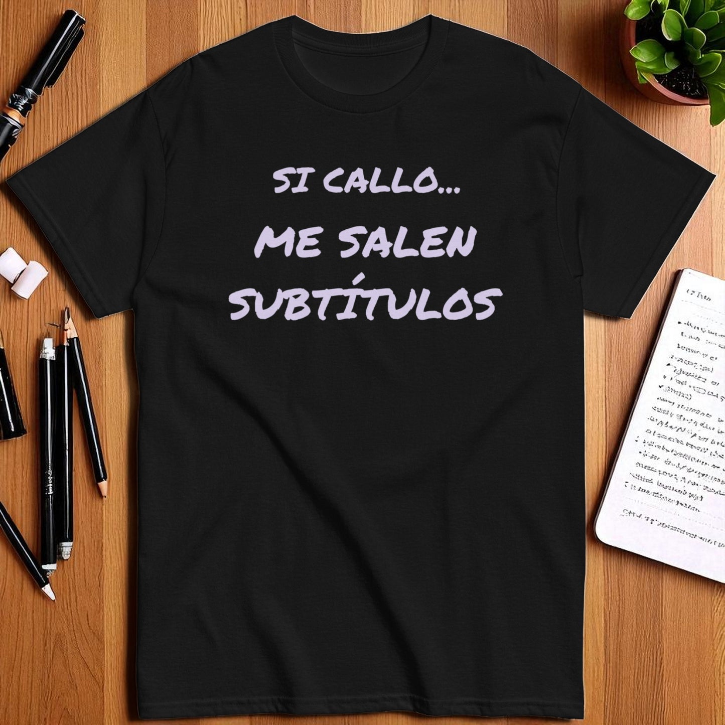 Camiseta negra unisex con frase Si callo me salen subtítulos en plano cenital con libreta y lápiz