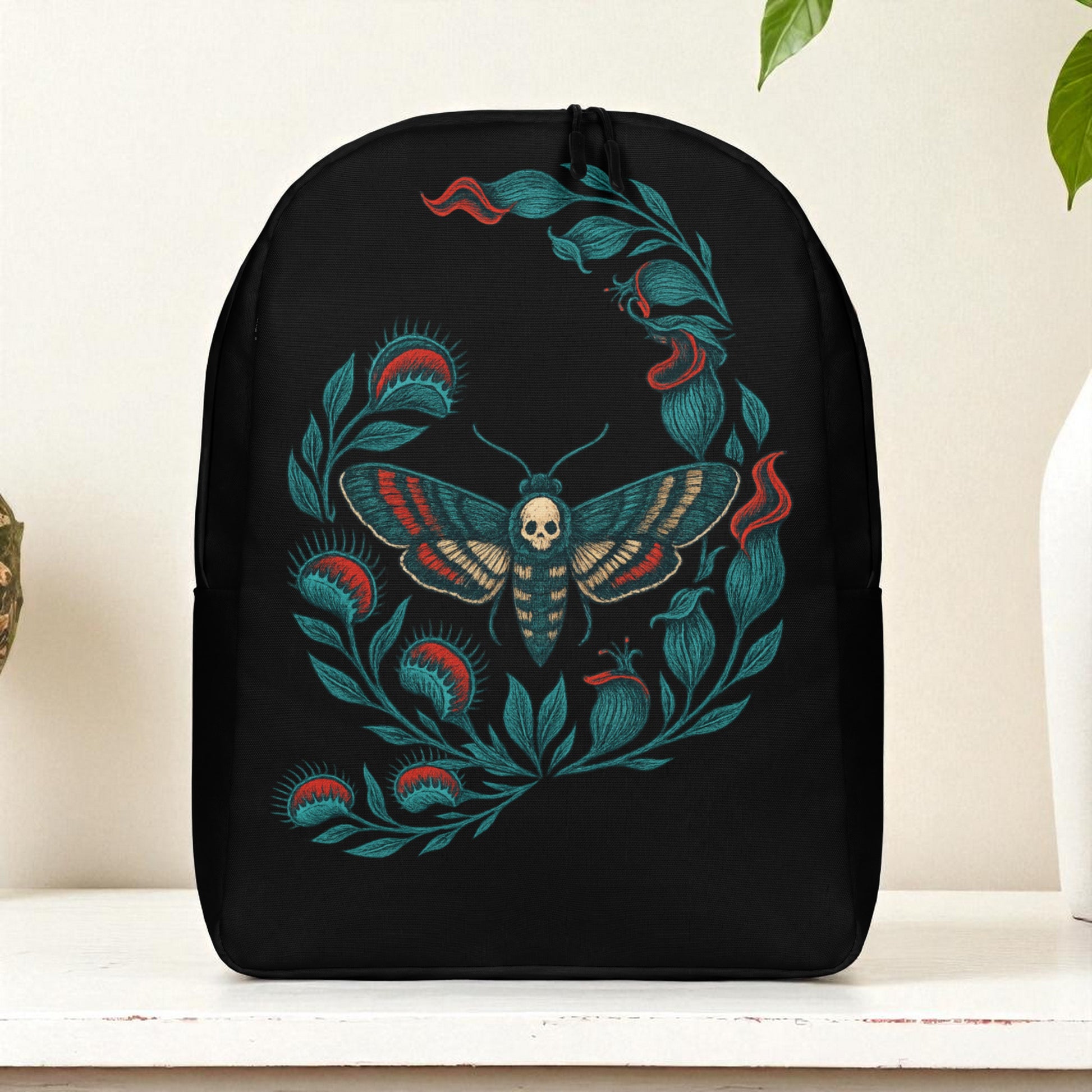 Mochila negra de frente sobre mesa, diseño de polilla calavera rodeada de hojas, fondo blanco con planta.