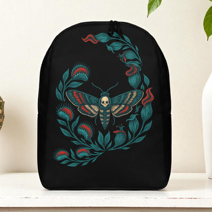 Mochila negra de frente sobre mesa, diseño de polilla calavera rodeada de hojas, fondo blanco con planta.
