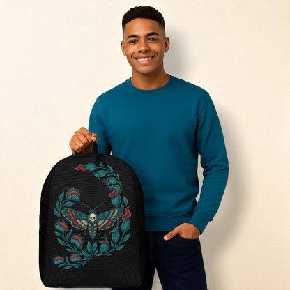 Hombre con camiseta azul petróleo sosteniendo mochila negra con polilla calavera, fondo blanco.