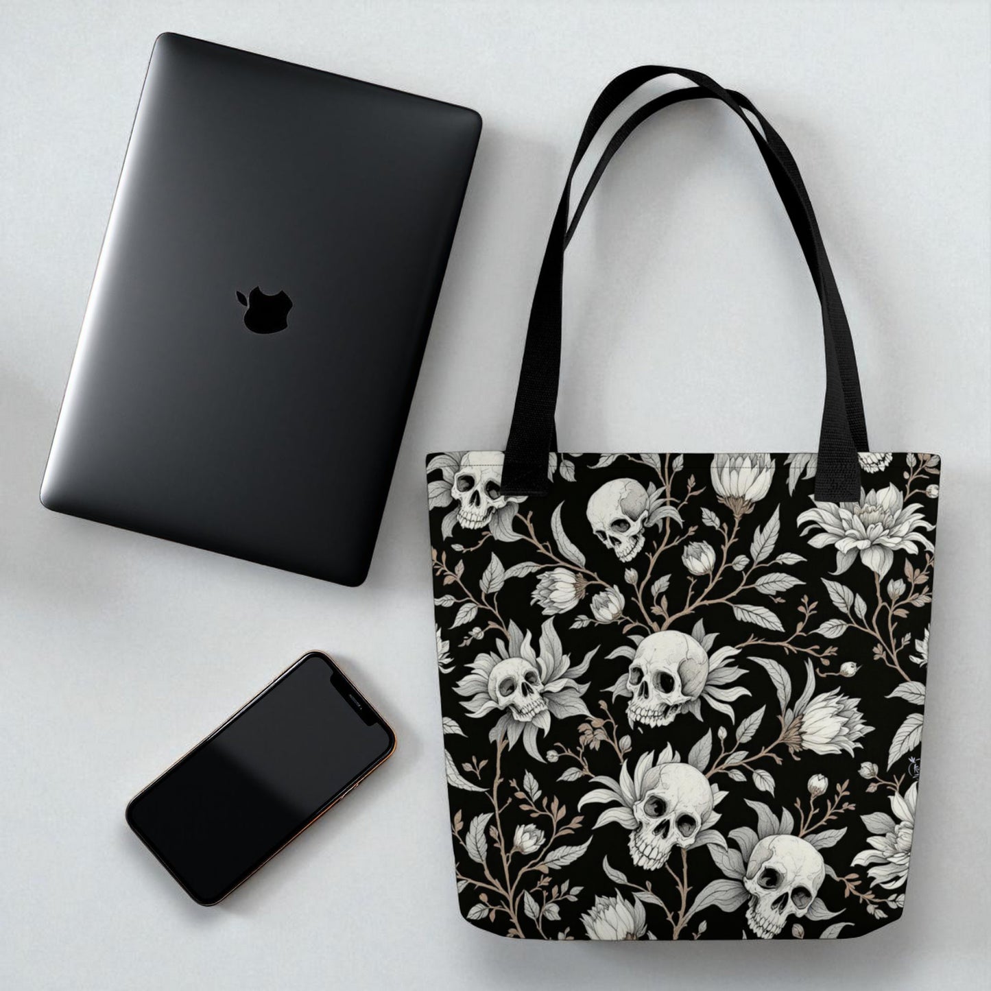 Bolso tote negro con calaveras y flores sobre mesa junto a portátil y móvil para referencia de tamaño