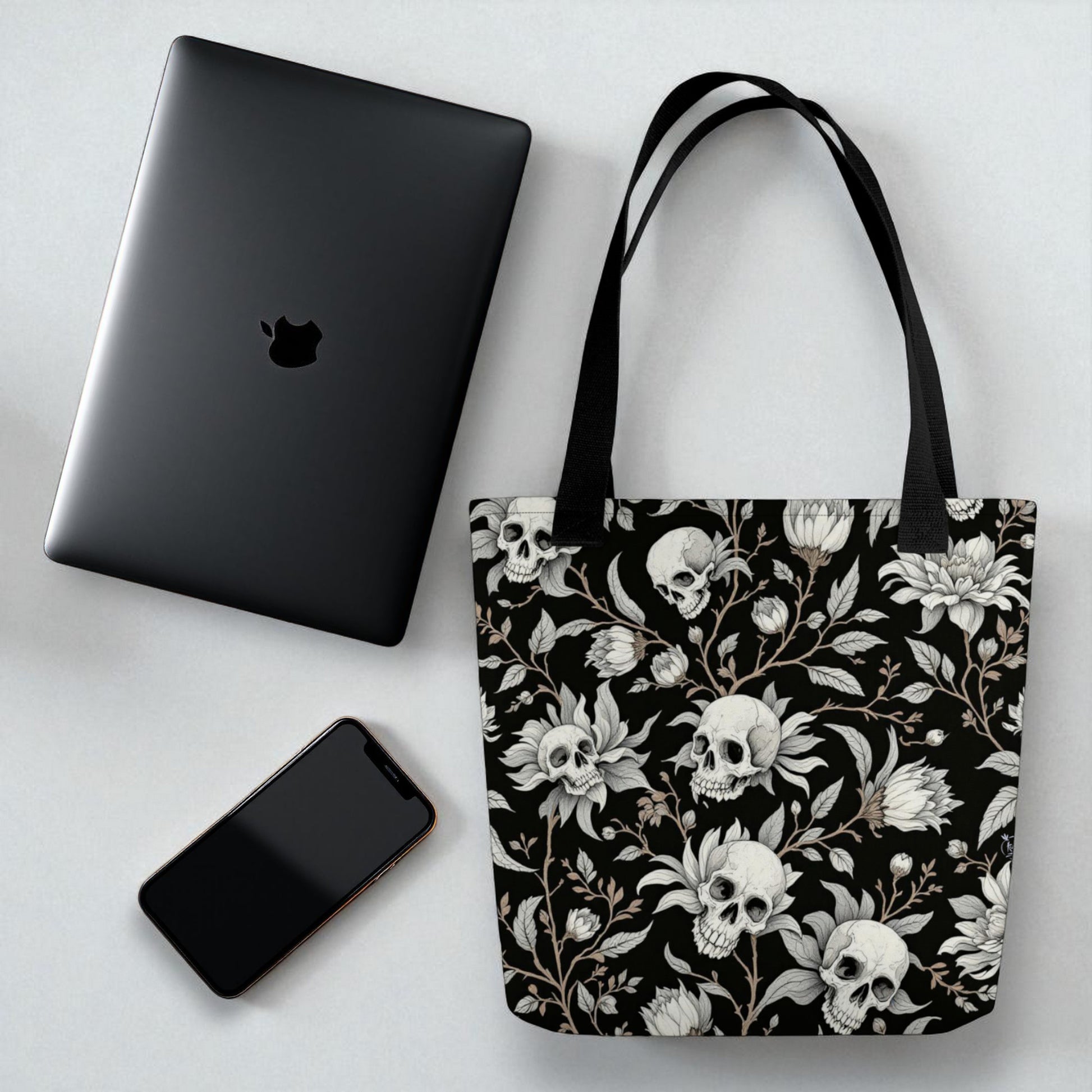 Bolso tote negro con calaveras y flores sobre mesa junto a portátil y móvil para referencia de tamaño
